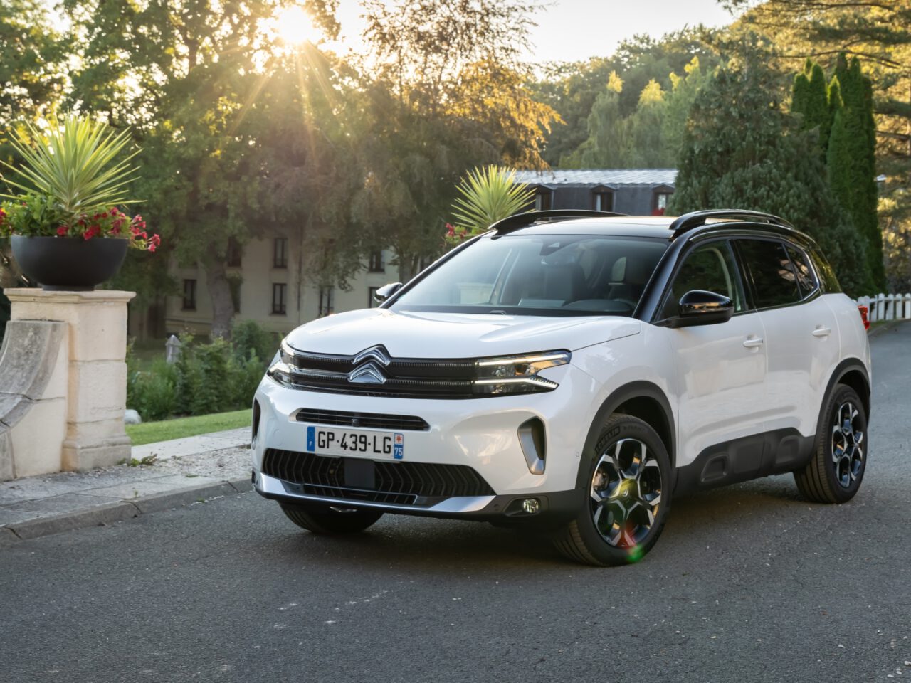 Een witte Citroën C5 Aircross SUV staat geparkeerd op een schilderachtige, met bomen omzoomde weg met een historisch gebouw op de achtergrond.