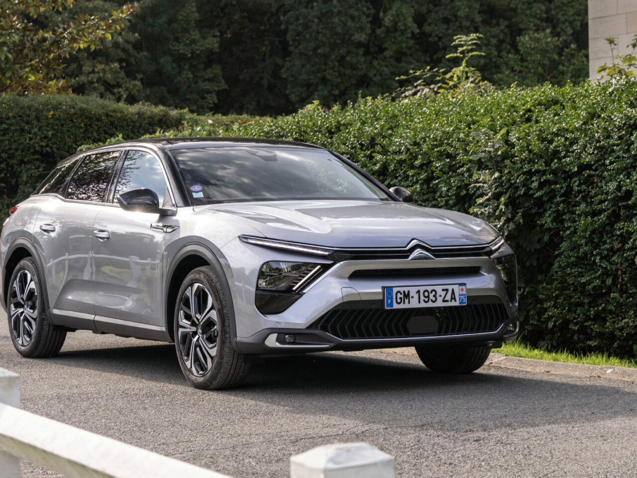 Een zilveren Citroën C5 X-auto staat geparkeerd op een weg, genesteld naast een weelderige groene heg en bomen. Op het kenteken van de auto staat "GM-193-ZA.