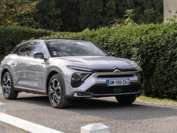 Een zilveren Citroën C5 X-auto staat geparkeerd op een weg, genesteld naast een weelderige groene heg en bomen. Op het kenteken van de auto staat "GM-193-ZA.