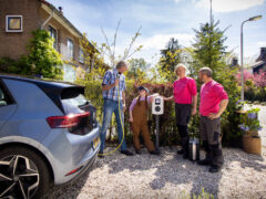 Vier mensen staan op een zonnige dag bij een elektrische auto die wordt opgeladen bij een privélaadpaal buiten een huis.