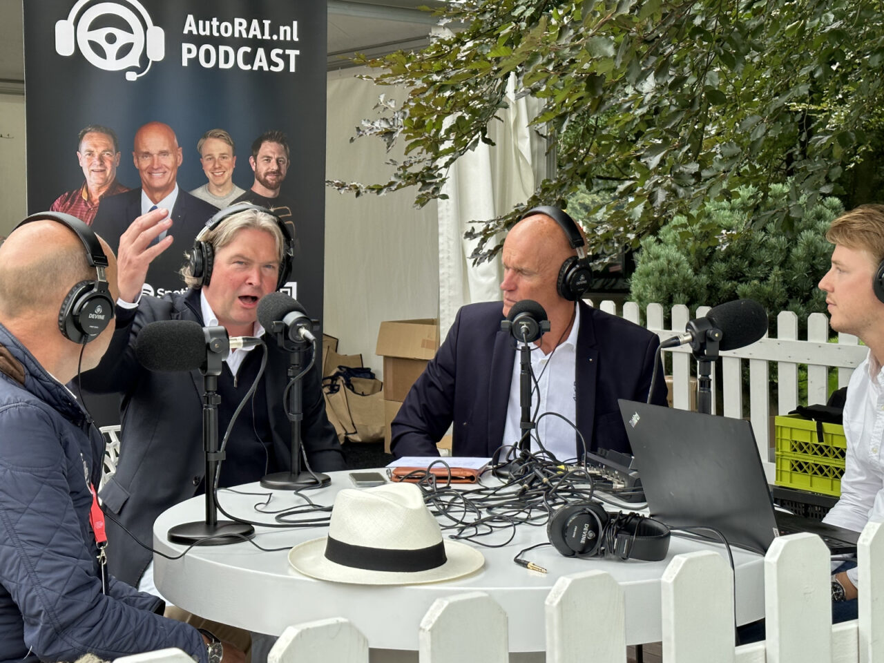 Vier mannen nemen een podcast op aan een ronde tafel. Microfoons, koptelefoons en een laptop zijn aanwezig. Op de achtergrond staat een promotieposter voor "AutoRAI Podcast #16".