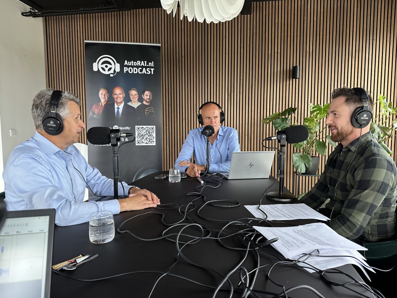 Drie mannen met een koptelefoon nemen AutoRAI Podcast #17 op. Ze zitten rond een tafel met microfoons, laptops en papieren. Op de achtergrond staat een podcast-promotieposter.