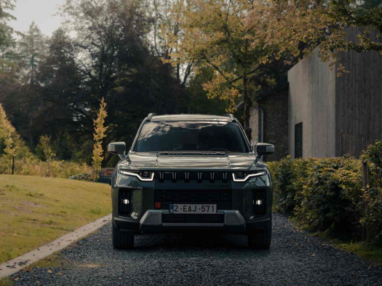 Een donkergekleurde SsangYong Torres SUV met moderne koplampen staat geparkeerd op een grindpad omgeven door bomen en groen, naast een gebouw met een houten buitenkant.