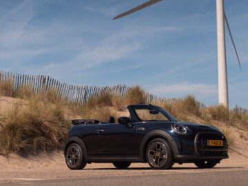 Een zwarte Mini Electric cabriolet geparkeerd op een zandweg nabij een houten hek en hoog gras, met een grote windturbine op de achtergrond onder een blauwe lucht.