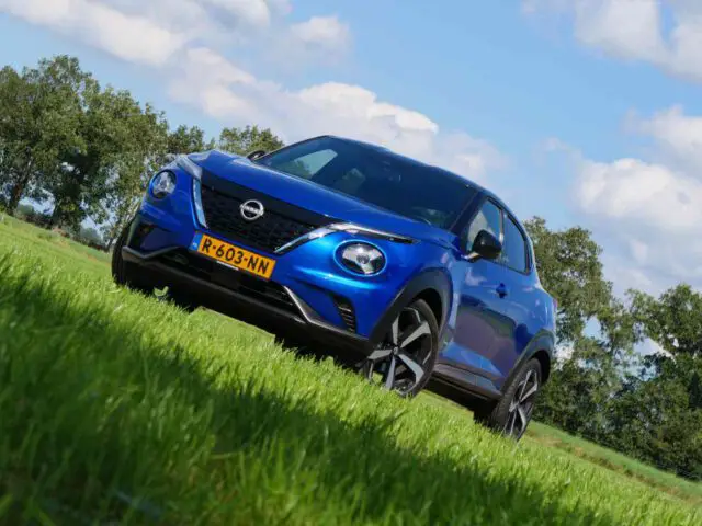 Een blauwe Juke hybride compacte SUV met Nederlands kenteken staat geparkeerd op een grasveld met bomen en een gedeeltelijk bewolkte lucht op de achtergrond.