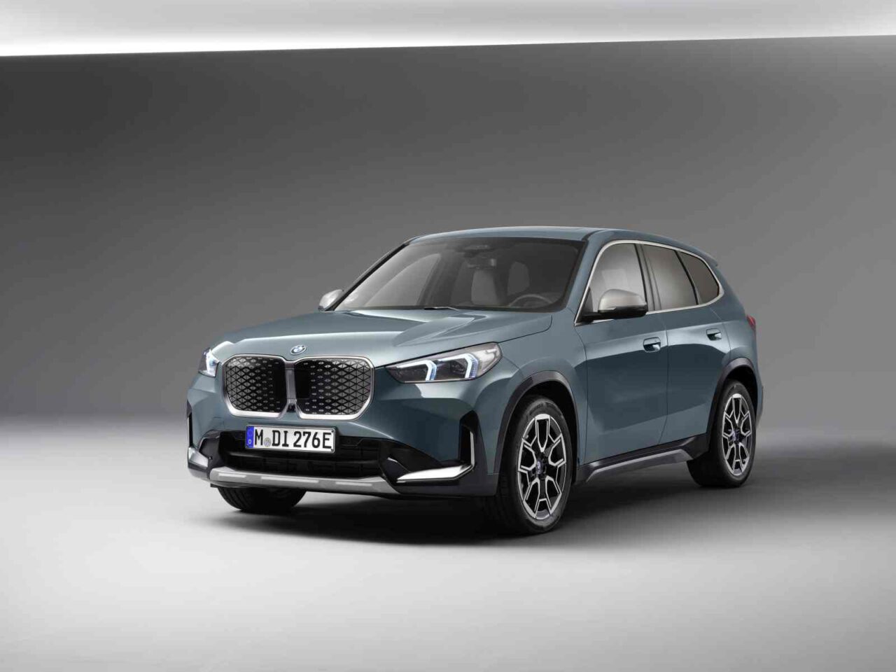 Een BMW ix1 SUV staat geparkeerd in een goed verlichte studio tegen een grijze achtergrond. Het blauwe voertuig laat zijn opvallende grille en moderne design naadloos zien.