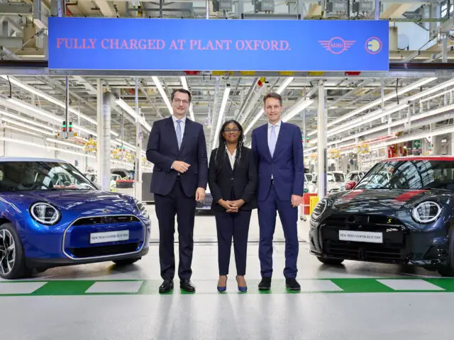 Drie mensen staan in een autoproductiefabriek tussen twee elektrische auto's, een blauwe en een zwarte. Op een bordje boven hen staat: 'Fully Charged at MINI Fabriek Oxford.