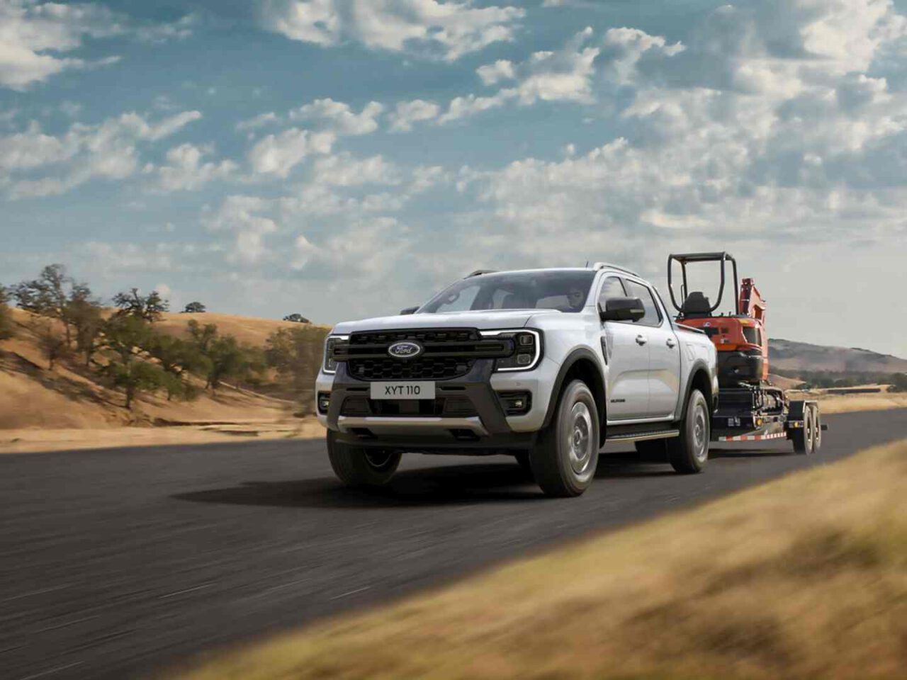 Een witte Ford pick-uptruck rijdt over een verharde weg en trekt een aanhangwagen met zware machines, met droge, glooiende heuvels en een gedeeltelijk bewolkte lucht op de achtergrond.