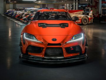 Een feloranje Toyota GR Supra GT4 100 Edition met agressieve styling en LED-koplampen wordt tentoongesteld in een moderne garage, omringd door andere raceauto's op de achtergrond.