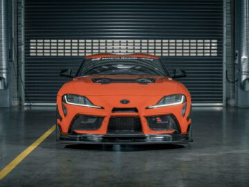 Vooraanzicht van een oranje Toyota GR Supra GT4 100 Edition racewagen in een garage, met een zwarte grille en een verlaagde bumper. De auto staat gecentreerd in de opname met een gedeeltelijk geopende sluiter op de achtergrond.