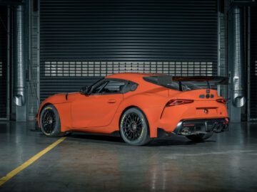 Een feloranje Toyota GR Supra GT4 100 Edition, voorzien van een grote achterspoiler, staat geparkeerd in een industriële garage met gesloten metalen luiken op de achtergrond.