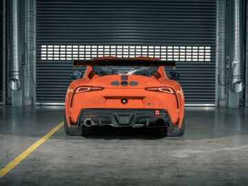 Achteraanzicht van een Toyota GR Supra GT4 100 Edition, met zijn levendige oranje tint en grote spoiler die opvalt terwijl hij geparkeerd staat in een industriële garage met gesloten rolluiken.