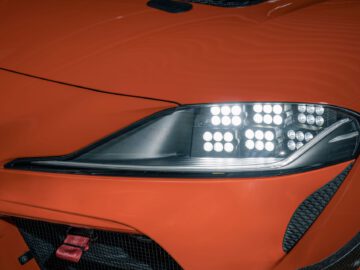 Close-up van de koplamp rechtsvoor van een oranje Toyota GR Supra GT4 100 Edition, met een unieke LED-lichtopstelling en scherpe lijnen in het ontwerp.