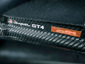 Close-upafbeelding van een Toyota GR Supra GT4 100 Edition-logo op een koolstofvezeloppervlak met een aangrenzend metalen plaatje met een auto-embleem en tekst.