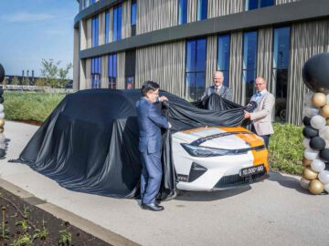 Drie mannen onthullen buiten een MG-auto bedekt met een zwarte stof, naast zwarte en gouden ballonnen, voor een modern gebouw.