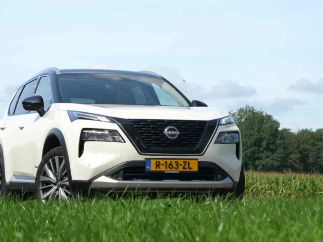 Een witte Nissan X-Trail E-Power met een geel kenteken staat geparkeerd op een grasveld, bomen op de achtergrond onder een heldere hemel.