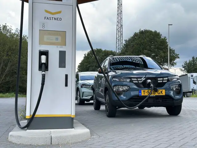 Onder een bewolkte hemel laden twee elektrische auto's, waaronder een Dacia Spring Electric, op bij een Fastned-station.