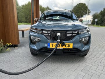 Een blauwe Dacia Spring Electric auto met Nederlands kenteken is aangesloten op een laadpaal op een verhard terrein, met bomen en een bewolkte lucht op de achtergrond.