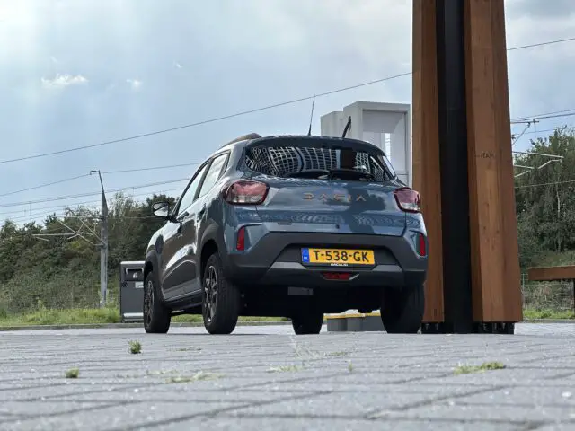 Een blauwe Dacia Spring Electric staat geparkeerd bij een laadstation op een verhard terrein met een grasachtige achtergrond. Op het kenteken van de auto staat "T-538-GK.