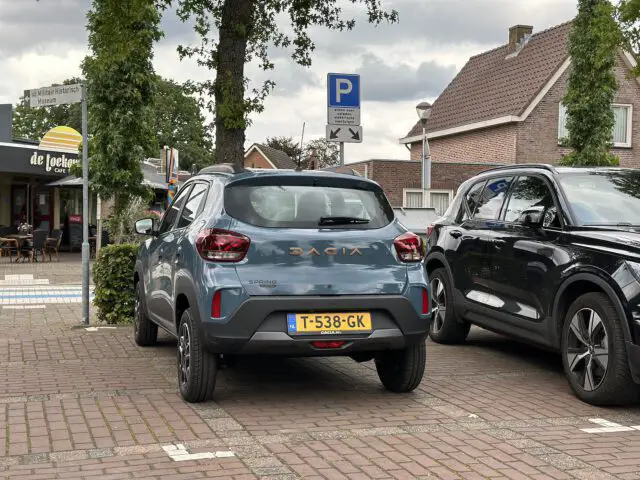 Een blauwe Dacia Spring Electric met Nederlands kenteken T-538-GK staat geparkeerd op een parkeerplaats naast een zwart voertuig, vlakbij een café en straatnaamborden.