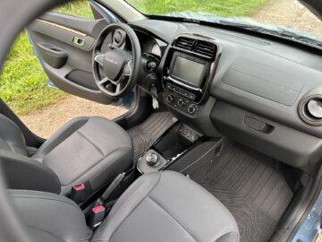 Het interieur van de Dacia Spring Electric toont de bestuurders- en voorpassagiersstoelen, een stuur en een dashboard met een leeg scherm. De versnellingspook en het bedieningspaneel zijn netjes gerangschikt, terwijl de autodeur aan de bestuurderszijde open staat.