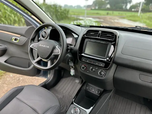 Binnenaanzicht van een Dacia Spring Electric met het stuur, het dashboard met touchscreen, bedieningsknoppen en een sleutel in het contact. Het interieur van de auto is uitgevoerd in een zwart en grijs kleurenschema.