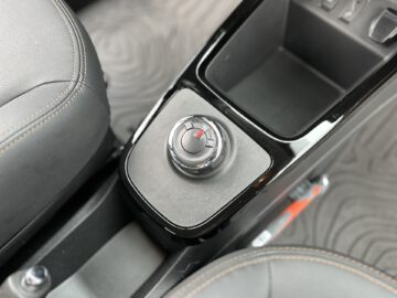 Close-up van de centrale bedieningsknop van een Dacia Spring Electric voor het aanpassen van instellingen, omgeven door leren en plastic onderdelen in het middenconsolegebied tussen twee stoelen.