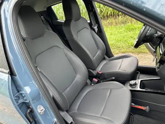 Het interieur van een blauwe Dacia Spring Electric, met de twee voorstoelen gemaakt van zwarte bekleding. Het bestuurdersportier staat open en de auto staat geparkeerd op een grasveld.