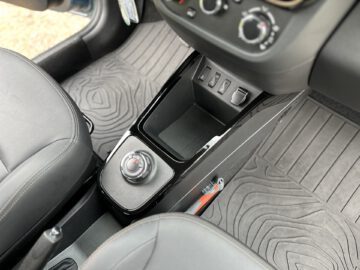 Close-up van de middenconsole van een Dacia Spring Electric met een draaiknop, verschillende knoppen, een klein opbergvak en rubberen vloermatten.