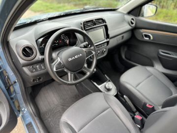 Een zicht op de bestuurders- en voorpassagiersstoelen van een Dacia Spring Electric-voertuig, met het dashboard, het stuur en de middenconsole met een aanraakscherm en verschillende bedieningselementen.