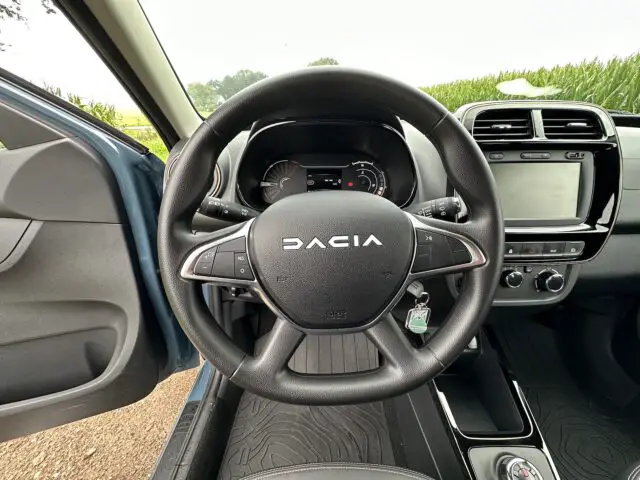 Het interieur van een Dacia Spring Electric-auto met het stuur, het dashboard en een deel van de bestuurdersdeur met het raam gedeeltelijk open.