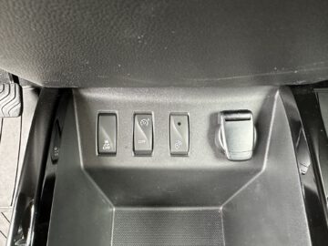 Close-up van een Dacia Spring Electric-bedieningspaneel met vijf knoppen: tractiecontrole uit, parkeersensor uit, onbekend symbool, nog een onbekend symbool en een draaiknop aan de rechterkant.