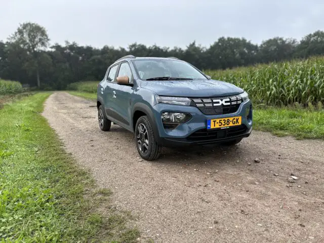 Een blauwe Dacia Spring Electric met Nederlands kenteken "T-538-GK" staat geparkeerd op een landelijke onverharde weg naast een veld met hoog maïs onder een bewolkte hemel.