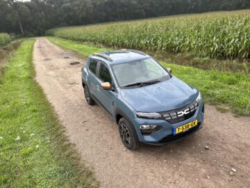 Een blauwe Dacia Spring Electric compacte SUV met een Europees kenteken staat geparkeerd op een onverharde weg naast een maïsveld.