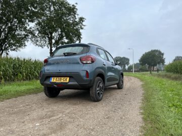 Een blauwe Dacia Spring Electric SUV geparkeerd op een onverharde weg naast een maïsveld, onder een bewolkte hemel. De auto heeft een Nederlands kenteken. Op de achtergrond staan bomen en een lantaarnpaal.