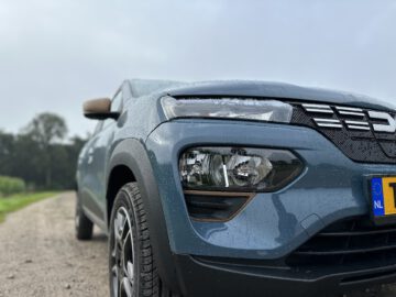 Close-up van de linkervoorkant van een blauwe Dacia Spring Electric, geparkeerd op een onverharde weg, met regendruppels zichtbaar op het voertuig. Aan de rechterkant is een gedeeltelijke weergave van het kenteken van de auto te zien.