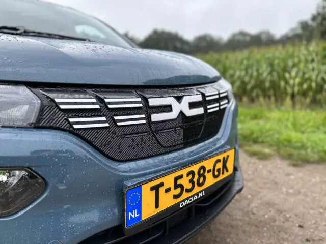 Close-up van de voorkant van een blauwe Dacia Spring Electric met een Nederlands kenteken met de tekst "T-538-GK". De auto is nat, mogelijk door de regen, en staat geparkeerd in de buurt van een grasveld met maïsvelden op de achtergrond.