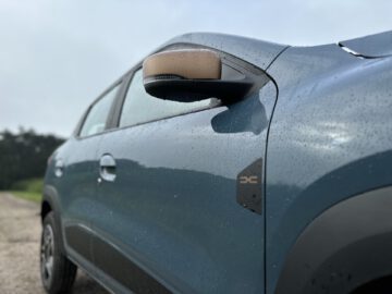 Close-up van de zijkant van een blauwe Dacia Spring Electric, met een met regendruppels bedekte zijspiegel en deur.