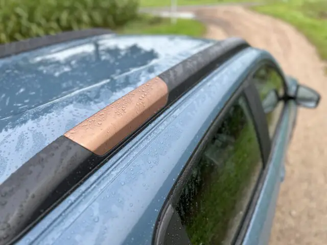 Close-up van een natte dakrail van een auto met regendruppels op een blauwe Dacia Spring Electric, geparkeerd naast een grasveld en een onverharde weg.