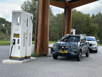 Twee elektrische voertuigen laden op bij een wegkantstation onder een houten overkapping. De voorste auto, een Dacia Spring Electric, wordt op de laadunit aangesloten, terwijl daarachter een ander voertuig geparkeerd staat.