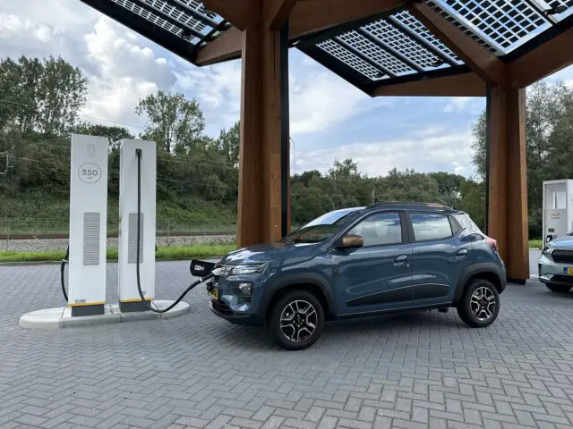 Een Dacia Spring Electric-voertuig wordt geparkeerd en opgeladen bij een station met een overdekt dak. Op de achtergrond zijn bomen en een bewolkte hemel zichtbaar.