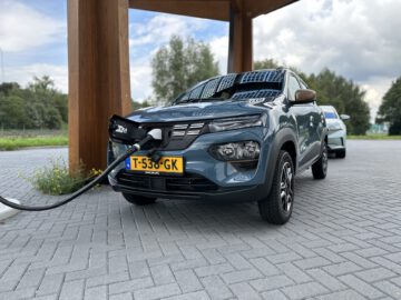 Een Dacia Spring Electric-auto wordt opgeladen bij een laadstation voor elektrische voertuigen, terwijl er direct achter een andere auto geparkeerd staat. Het tafereel ontvouwt zich buiten onder een houten structuur.