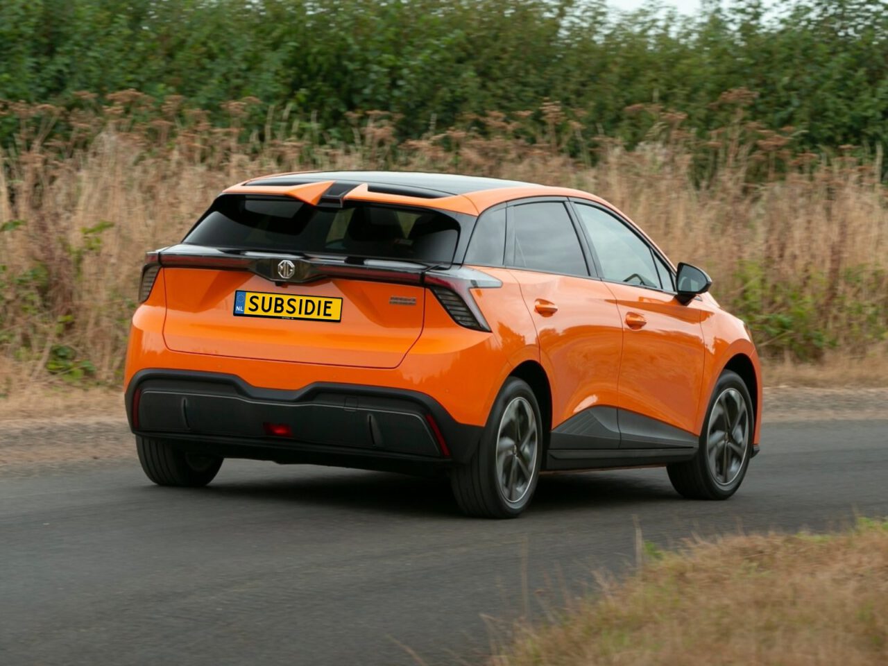 Een oranje elektrische auto uit China met het kenteken "SUBSIDIE" rijdt op een verharde weg, met droog gras en struiken langs de kant van de weg.