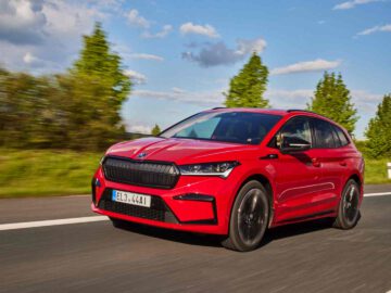 Een rode Skoda Enyaq compacte SUV rijdt op een weg met groen en bomen op de achtergrond onder een gedeeltelijk bewolkte hemel. Op het kenteken staat "EL3 4441.