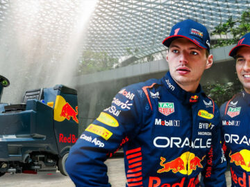 Twee personen in race-uniformen, mogelijk Verstappen-fans, staan voor een grote installatie op de Jewel Changi Airport in Singapore. De scène toont een betoverende waterval en een machine die prominent op de voorgrond staat.
