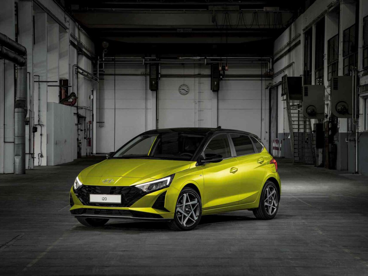 Een gele Hyundai i20 hatchback staat geparkeerd in een industrieel pakhuis met hoge plafonds en grote deuren op de achtergrond.