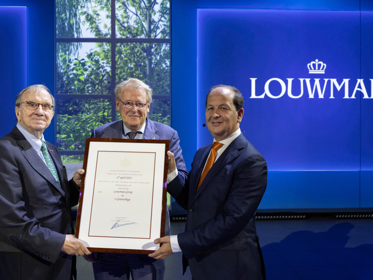 Drie mannen in pak staan op een podium voor een blauwe achtergrond met het woord 'LOUWMAN'. Twee van de mannen houden een ingelijst certificaat omhoog, ter ere van het behalen van het prestigieuze Predicaat Koninklijk door de Louwman Groep.