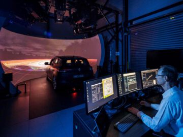 Een persoon bedient meerdere computermonitors terwijl hij een Renault-voertuig test in een geavanceerde rijsimulatieopstelling met een gebogen scherm dat een virtuele omgeving in een donkere kamer weergeeft.