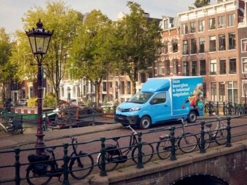 Een lichtblauwe Toyota-bestelauto staat geparkeerd in een smal straatje met een gracht, fietsen en historische gebouwen op de achtergrond.