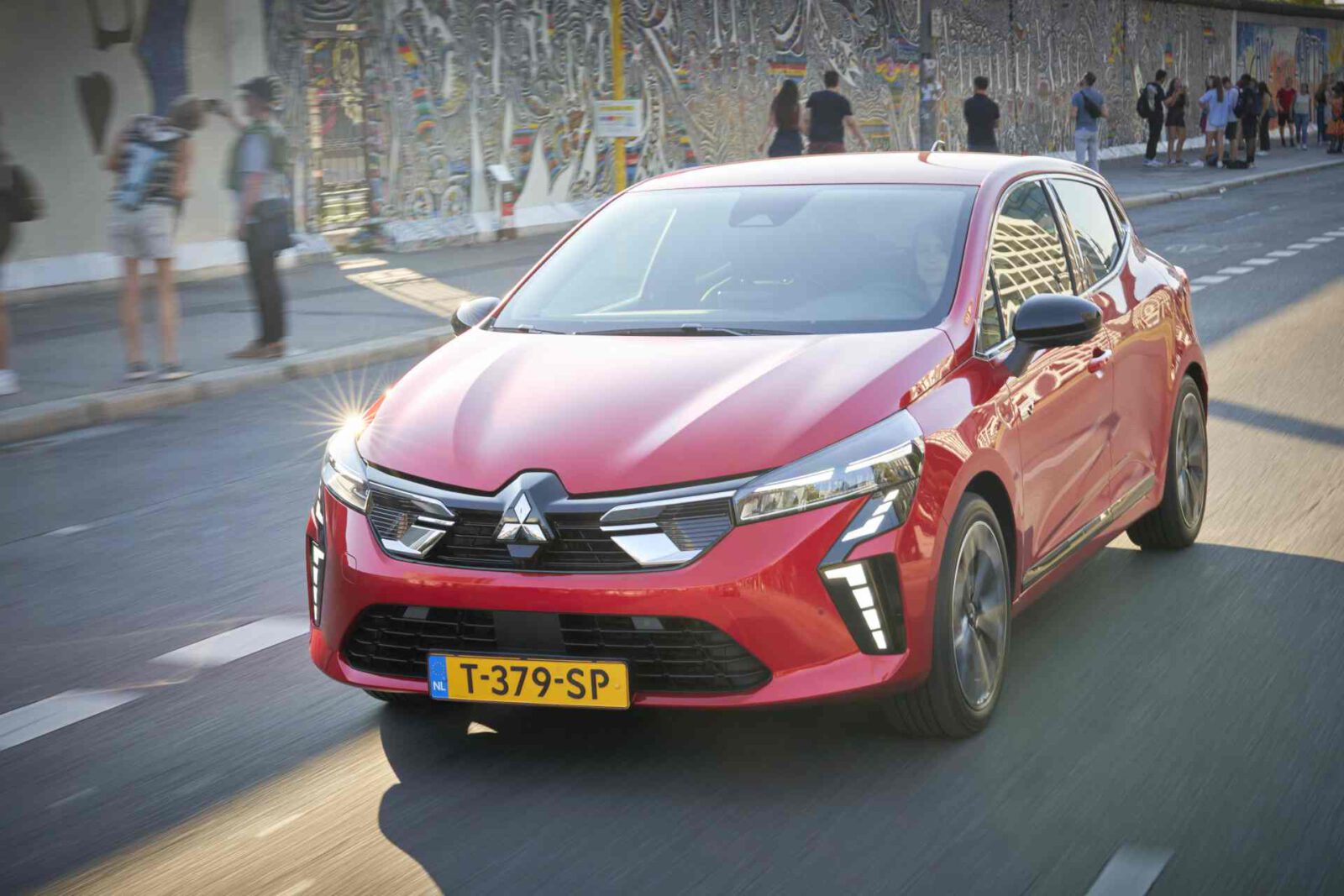 Een rode Mitsubishi Colt met een geel kenteken rijdt door een stadsstraat met een met muurschildering bedekte muur op de achtergrond.